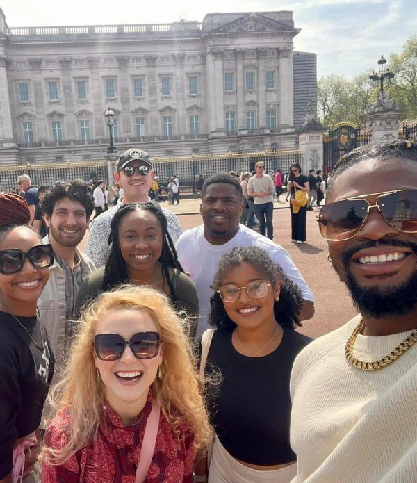 A group of young people pose for a selfie at Buckingham Palace. It is a bright sunny day. 