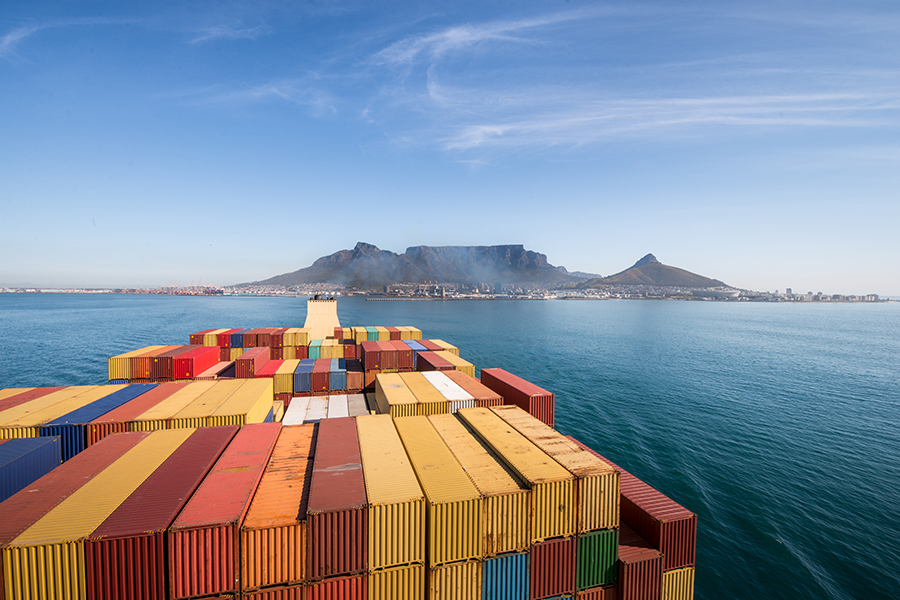 A large stacked container ship sailing on the ocean. A mountain and city are in the background.