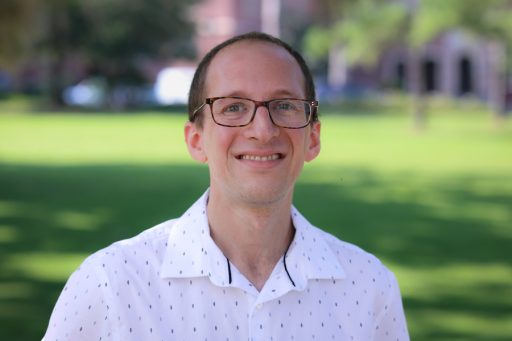 A smiling man in front of a sunny, grassy clearing