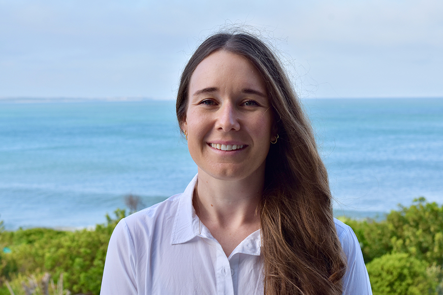 A woman smiling in front of a background of foliage and the ocean.