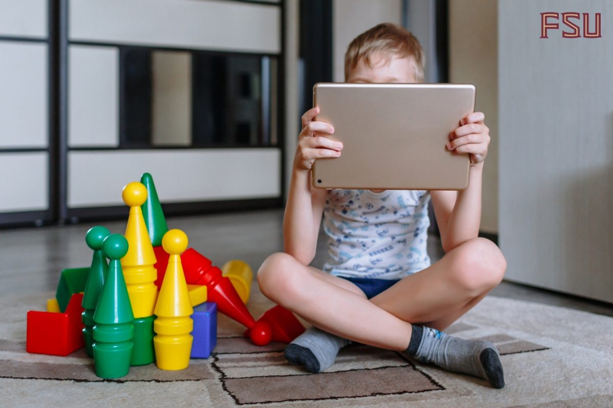 A young boy sits next to toys while looking at a tablet.
