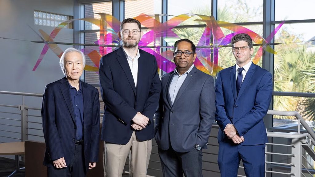 Four men in professional business attire stand in front of an abstract sculpture and large windows.