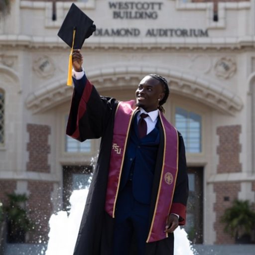 Photo of McKenzie Simon in his graduation gown, holding up his graduate cap in front of the Westcott building.