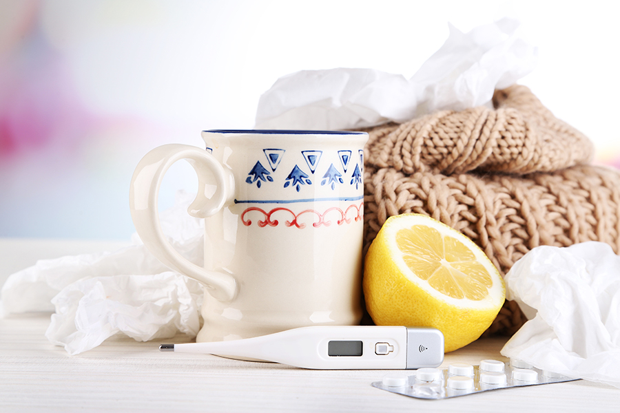 Photo of a mug next to a lemon, a thermometer, and medicine, in front of tissues and a blanket.