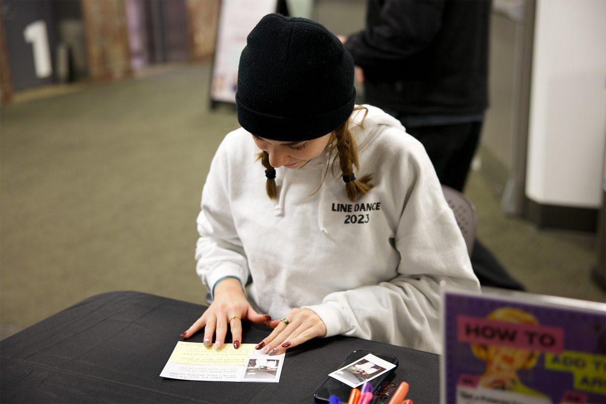 A student sits at a table filling out a note card for the “Add to the Archive at Strozier” event, with a Polaroid photo and markers placed in front of them.