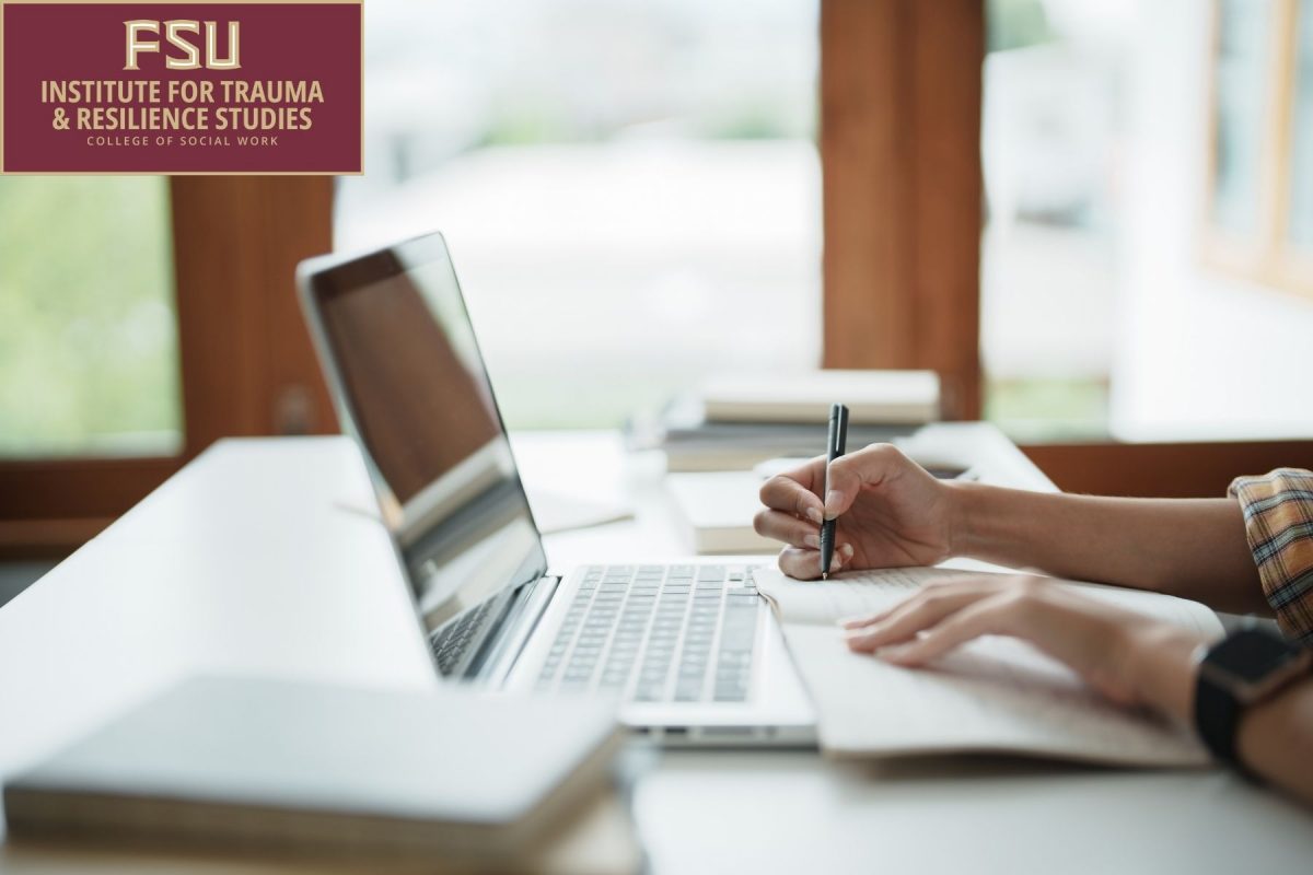 A person takes handwritten notes in a notebook while working on a silver laptop at a bright, sunlit desk, with the Florida State University Institute for Trauma & Resilience Studies logo in the top-left corner.