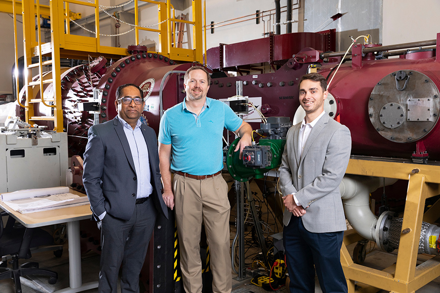 Three men stand in front of engineering equipment.