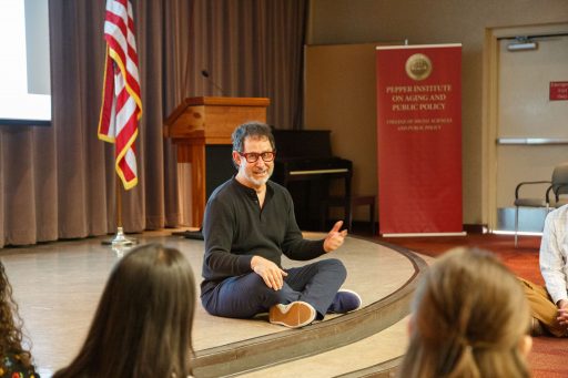 A man sits cross-legged on a stage in front of an audience, also sitting cross-legged.