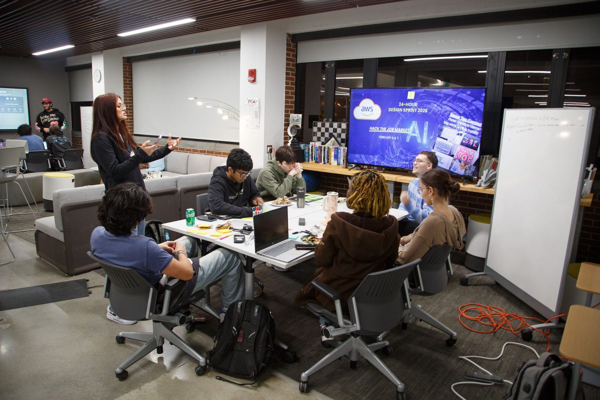 Students sit at a table with their computers, listening to someone standing speak to them.