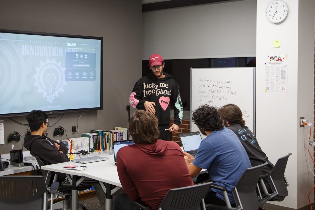STudents sit at a table and discuss, one of them standing and talking to the rest.