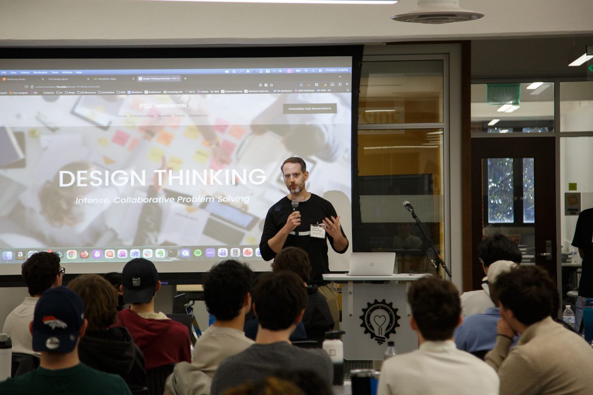 Students sit at tables watching and listening to someone speaking in front of them by a presentation.
