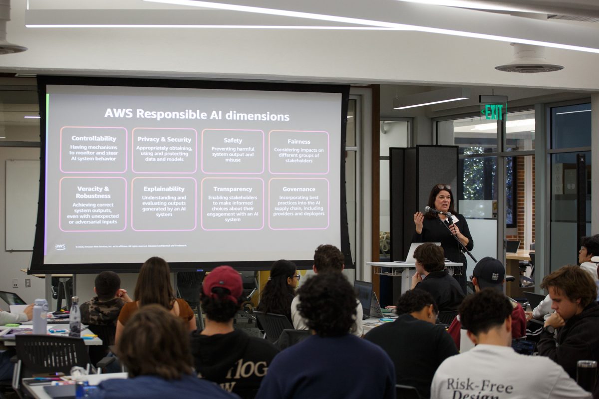 Students sit at tables watching and listening to someone speaking in front of them by a presentation.