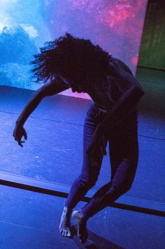 A dynamic shot of a dancer in motion, leaning low toward the stage floor. The lighting is moody and cool-toned, highlighting the silhouette of the performer against a colorful, ethereal backdrop projection.