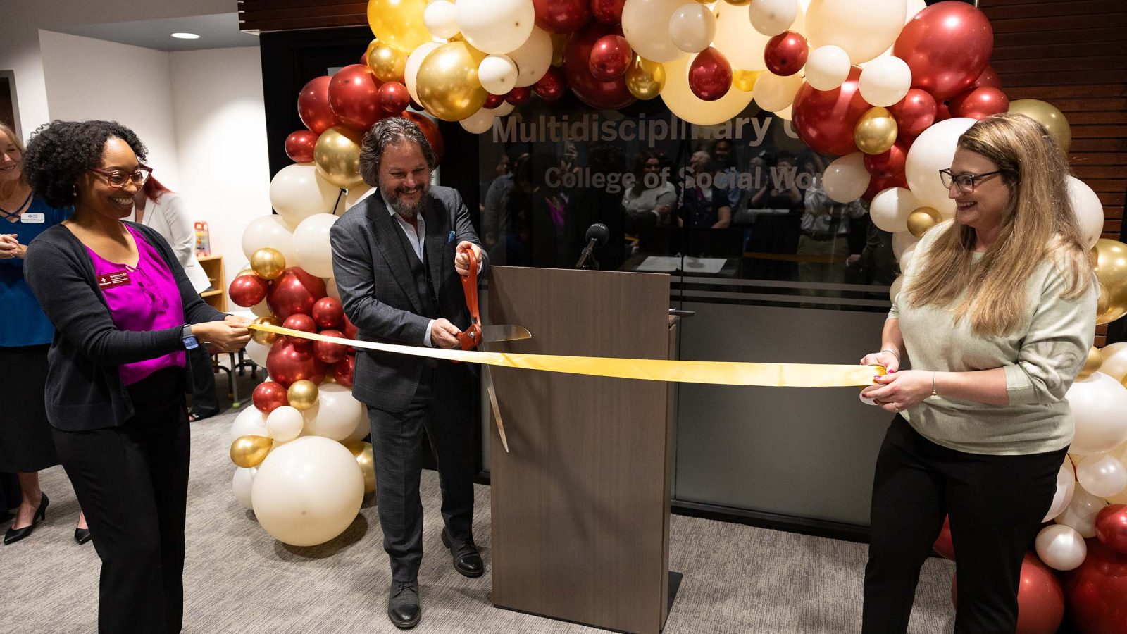 Two women hold either ends of a ribbon while a man starts to cut it with big scissors.