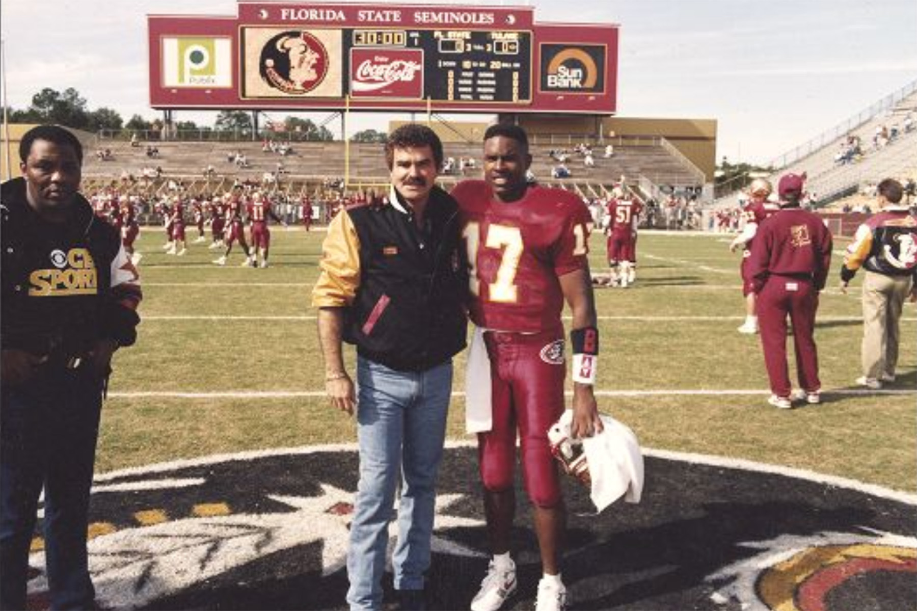 A vintage photograph of actor Burt Reynolds and FSU football player Charlie Ward standing at midfield in Doak Campbell Stadium. Reynolds wears a team jacket and Ward is in his garnet and gold jersey (number 17). The stadium scoreboard is visible in the background.