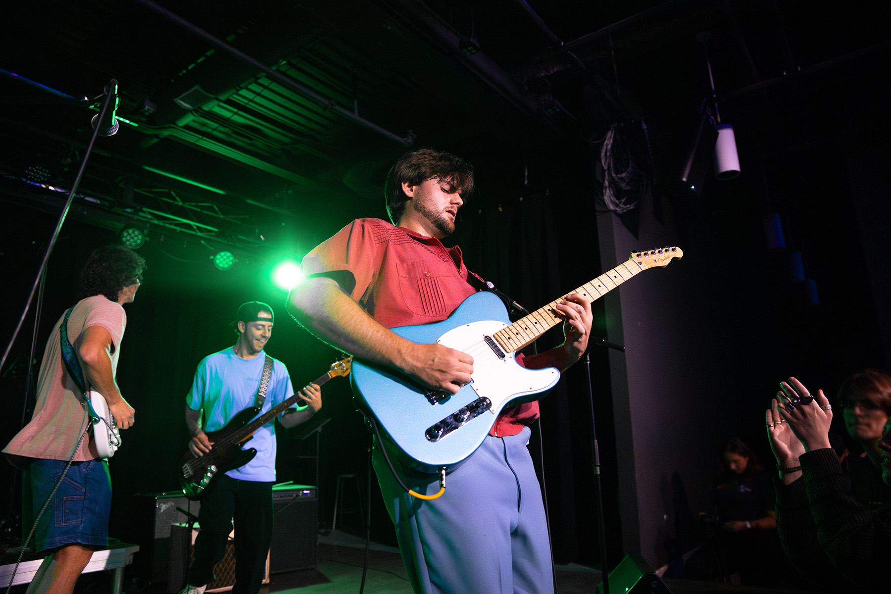 A band performs on a dark stage under a vibrant green spotlight, featuring a guitarist in a red shirt playing a metallic blue electric guitar. The scene captures an energetic live show with a bassist and an applauding audience member visible in the background.