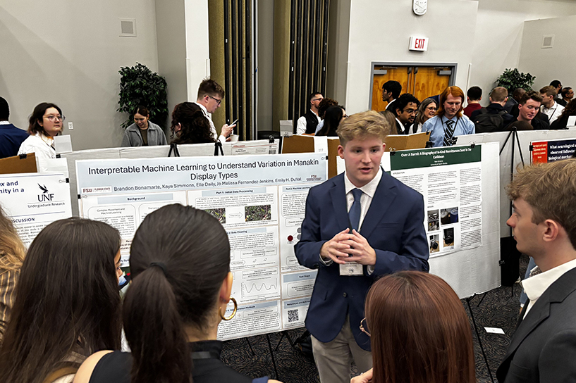 A man in a blue suit jacket speaks in front of a research poster.