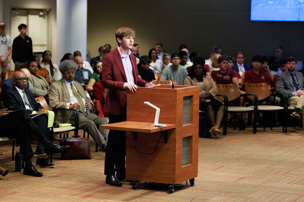A person stands at a wooden podium addressing a public meeting, with seated attendees listening in rows behind them in a large civic meeting room.