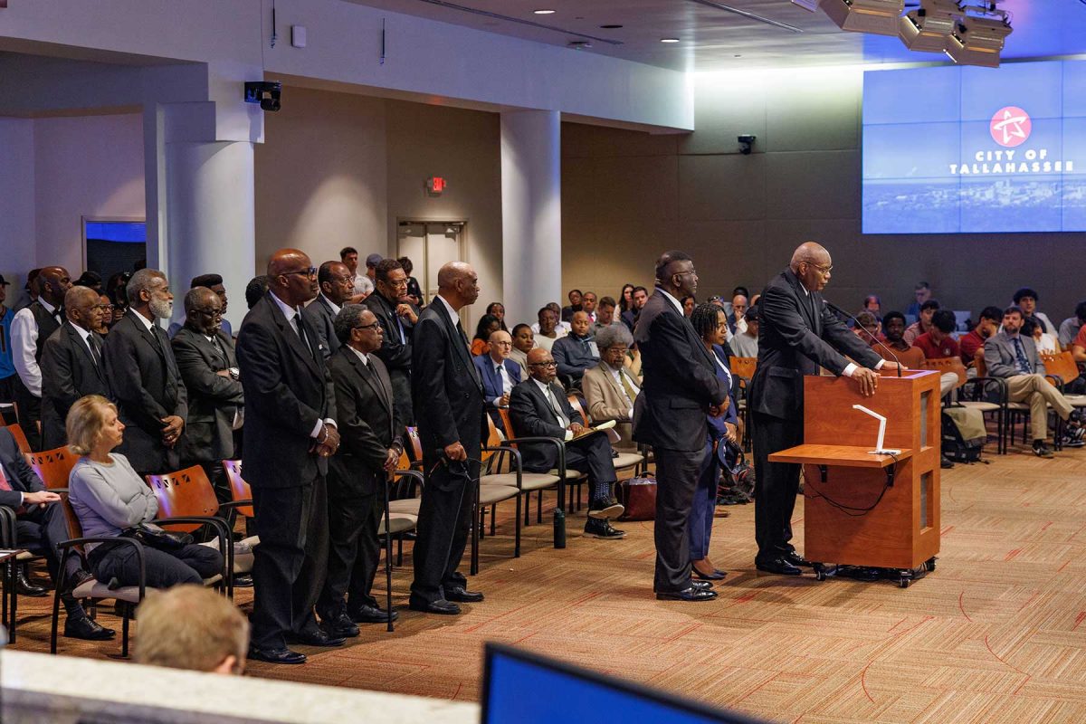 A speaker stands at a podium addressing a large audience in a City of Tallahassee commission chamber, while a line of formally dressed attendees stands nearby and others sit listening throughout the room.