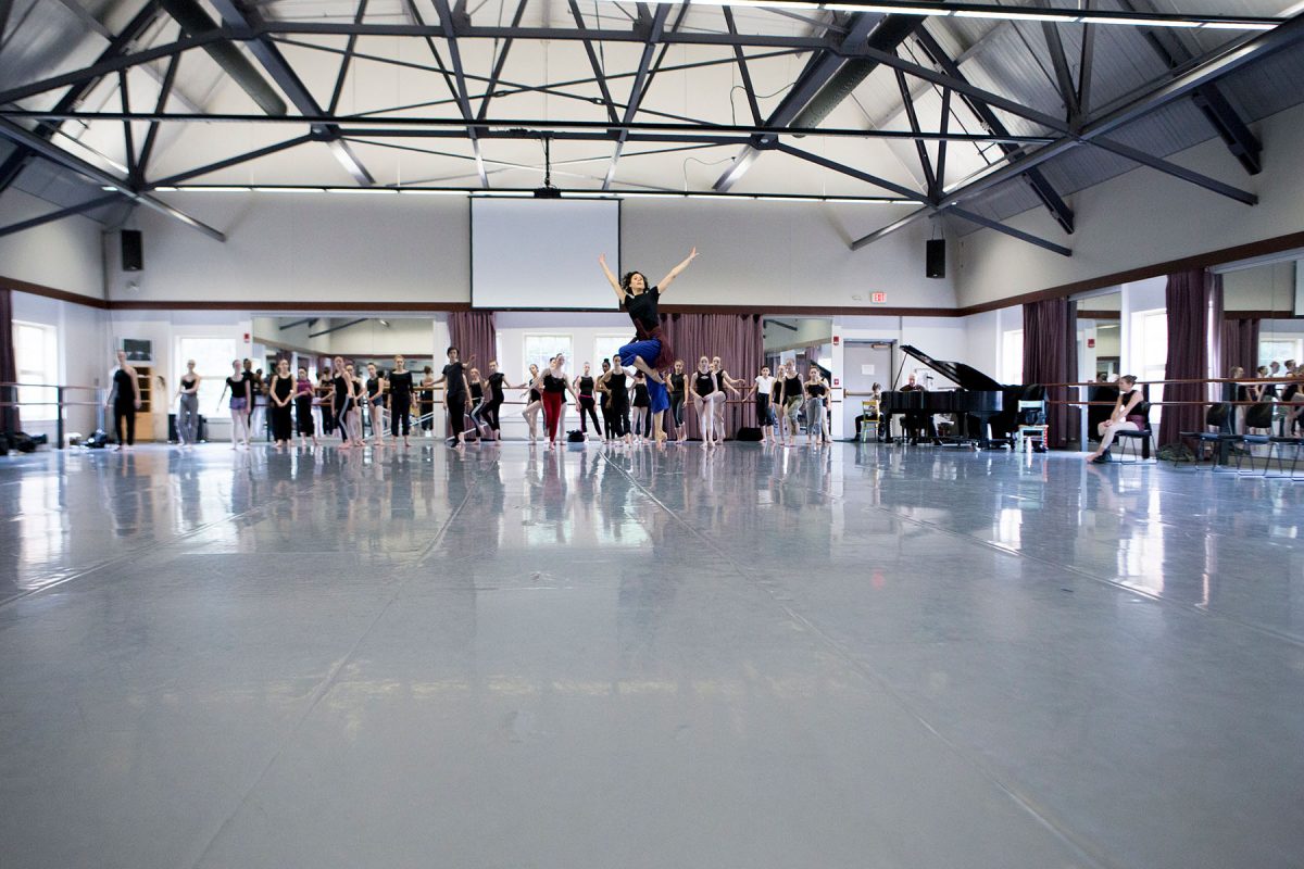The image captures a wide-angle, low-perspective view of a large, high-ceilinged dance studio with a group of dancers.