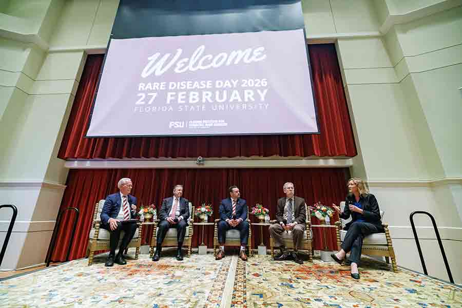 A group of five professionals—David Ledbetter, Richard McCullough, Adam Anderson, Eric Green, and Sarah South—stand in a row at the front of a room at the FSU College of Medicine. They are addressing an audience from a stage or podium area in observance of Rare Disease Day.