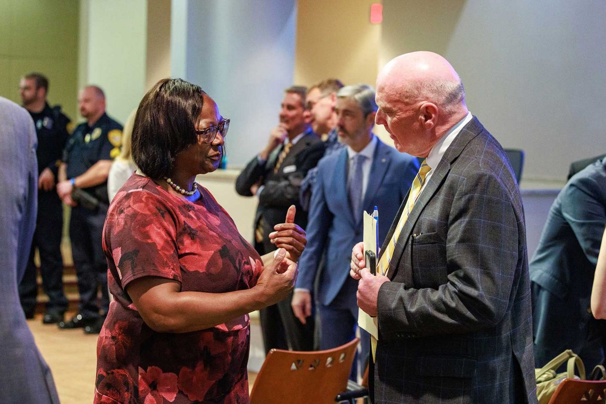 Two people stand and converse in a civic meeting space, one gesturing while the other holds documents, with additional attendees and uniformed officers visible in the background.