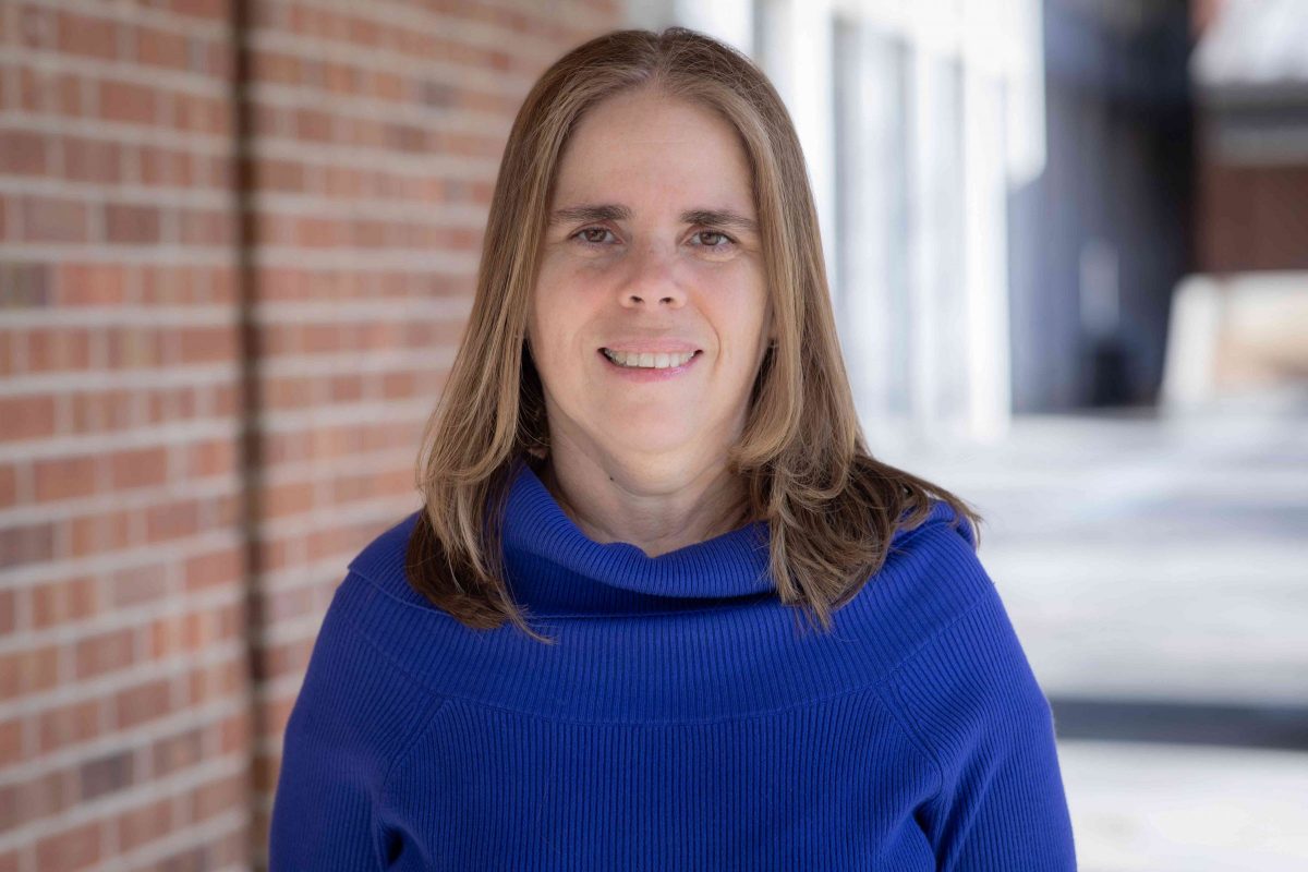 Headshot of a woman with light brown hair smiling with a brick building in the backdrop.