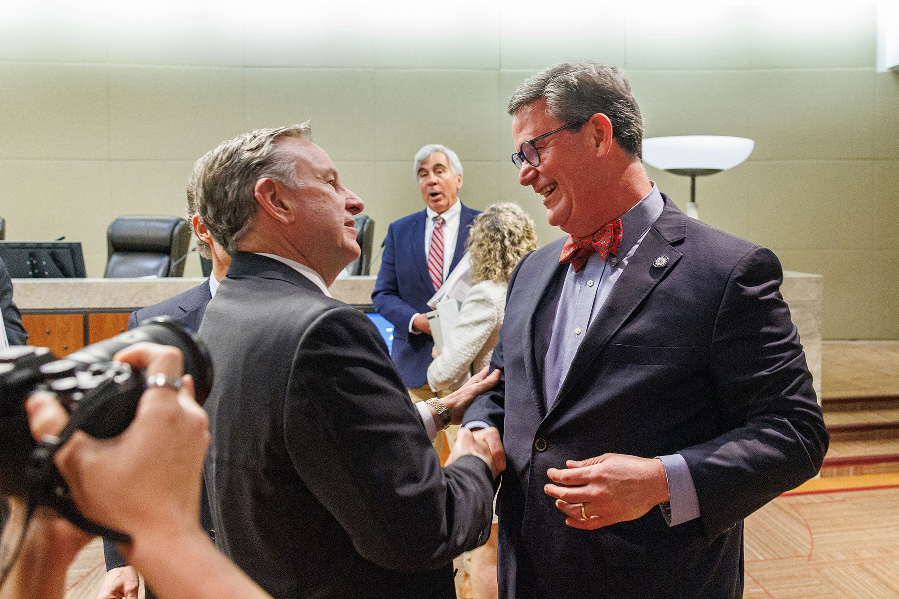 Two men in business suits greet each other and shake hands inside a government meeting chamber, with other attendees standing in the background and a photographer capturing the moment in the foreground.