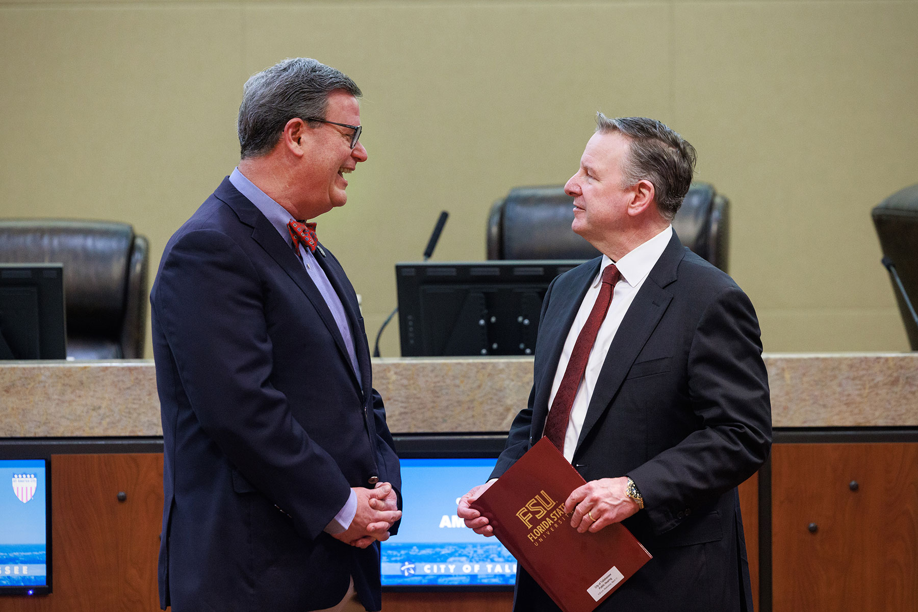 Two men in business suits stand facing each other in a formal council chamber, appearing to converse. One holds a maroon folder labeled “FSU Foundation,” with a dais, chairs, and digital screens visible in the background.