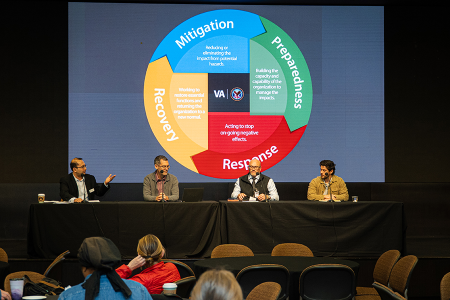 Four people sit at a table facing an audience for a discussion. Behind them is a logo that reads 