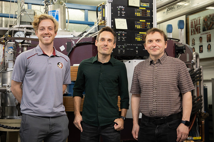 Three people stand in front of equipment in a physics laboratory.