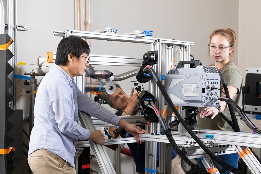 Three researchers work together on a complex experimental rig, adjusting sensors and structural components while cables run across the frame in a laboratory environment.