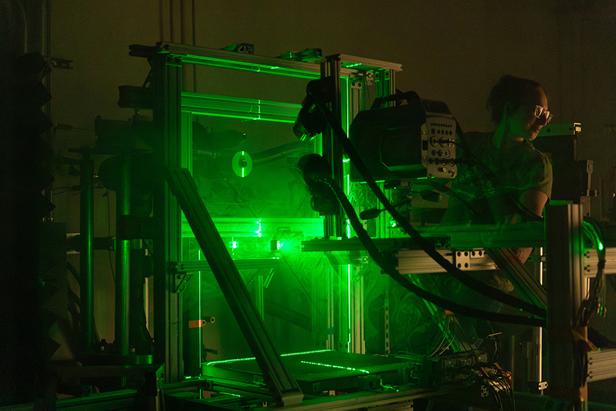 A person wearing glasses stands beside a laser-illuminated test rig, monitoring equipment with green light outlining the structure and internal components.