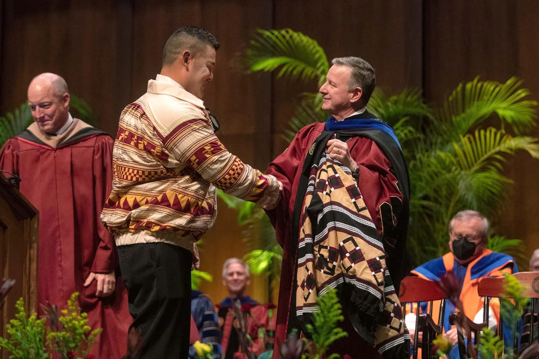 A man in a patterned patchwork jacket shakes hands with a university official in graduation regalia.