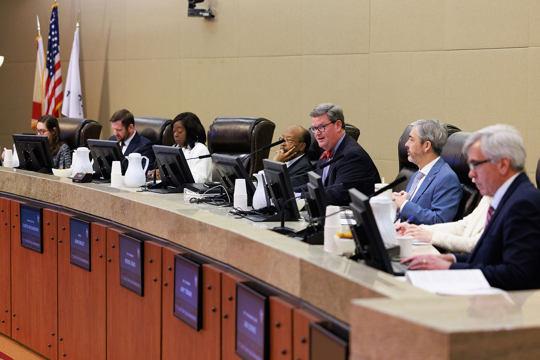 A panel of officials sits behind a long dais in a formal government meeting room, reviewing materials on computer monitors, with flags displayed behind them and nameplates along the front of the desk.