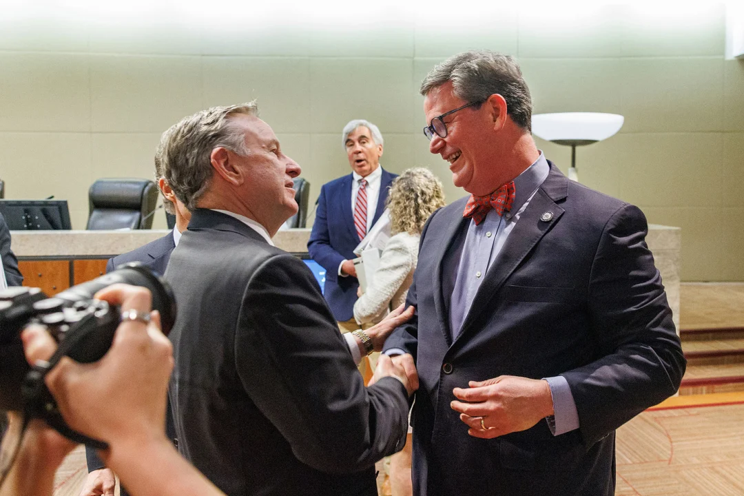 Two men in business suits greet each other and shake hands inside a government meeting chamber, with other attendees standing in the background and a photographer capturing the moment in the foreground.