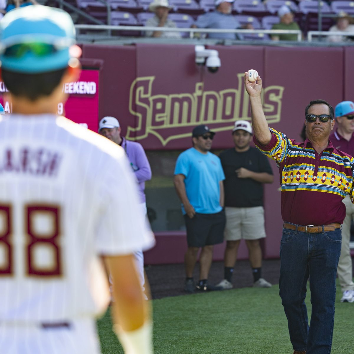 A man on a baseball field is in action as he starts to throw a baseball to a baseball player who is blurred out, with people in the background