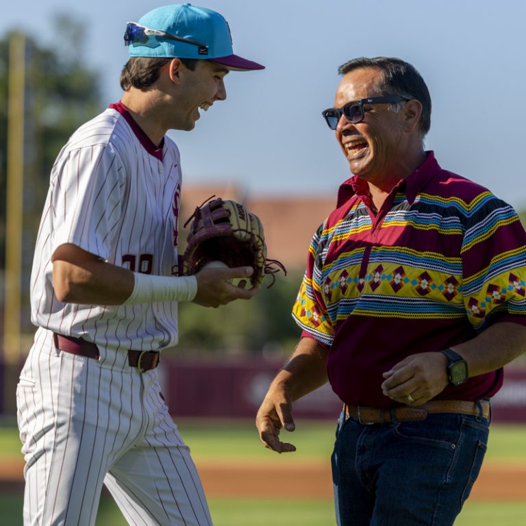 A baseball player holding a ball and a glove laughs with a man standing next to him