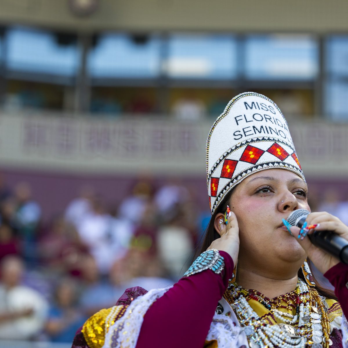 A woman wearing traditional indigenous clothing sings with people in a stadium blurred in the background