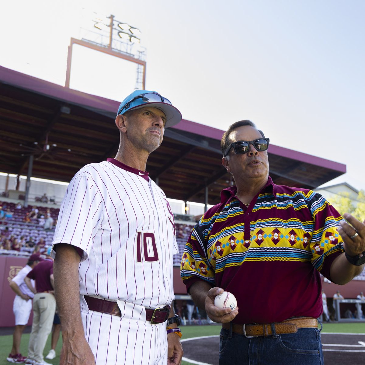Two men stand and talk at a baseball field, one wearing a baseball uniform and the other holds a baseball
