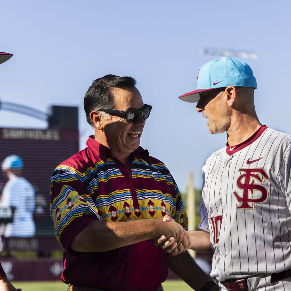 Two men shake hands at a baseball field, one wearing a baseball uniform