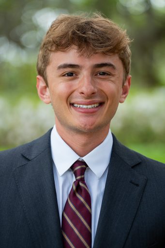 A young man with light brown hair wearing a dark grey suit, a white dress shirt, and a striped maroon and navy tie.