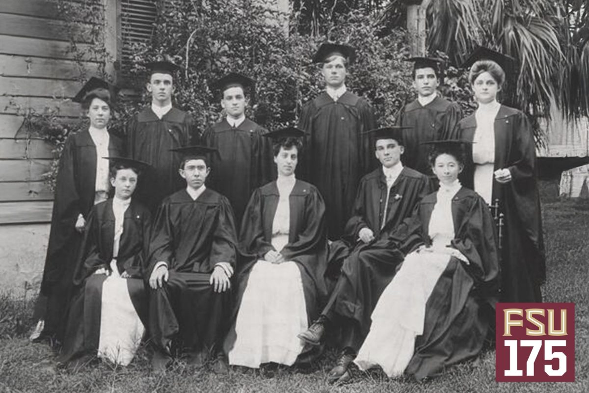 A group of eleven graduates in traditional academic caps and gowns poses for a formal black-and-white class portrait outdoors in front of a wooden building and greenery.