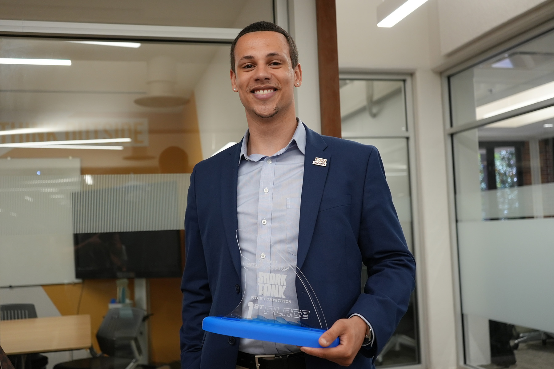 A smiling man in a blue blazer and light blue button-down shirt proudly displays a clear glass "Shark Tank Pitch Competition 1st Place" trophy with a blue base in a modern office environment.