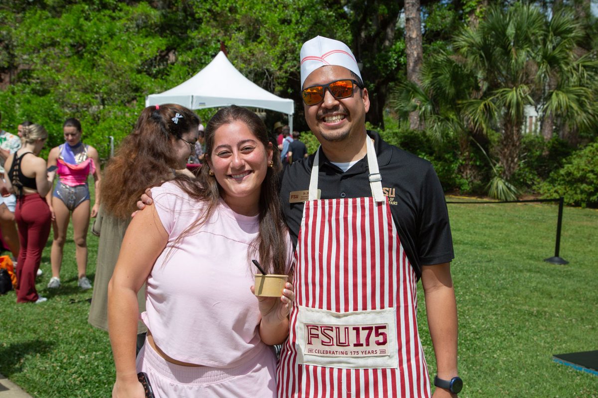 Two people outside smile, one wearing a red and white apron with a hat, the other holding a cup