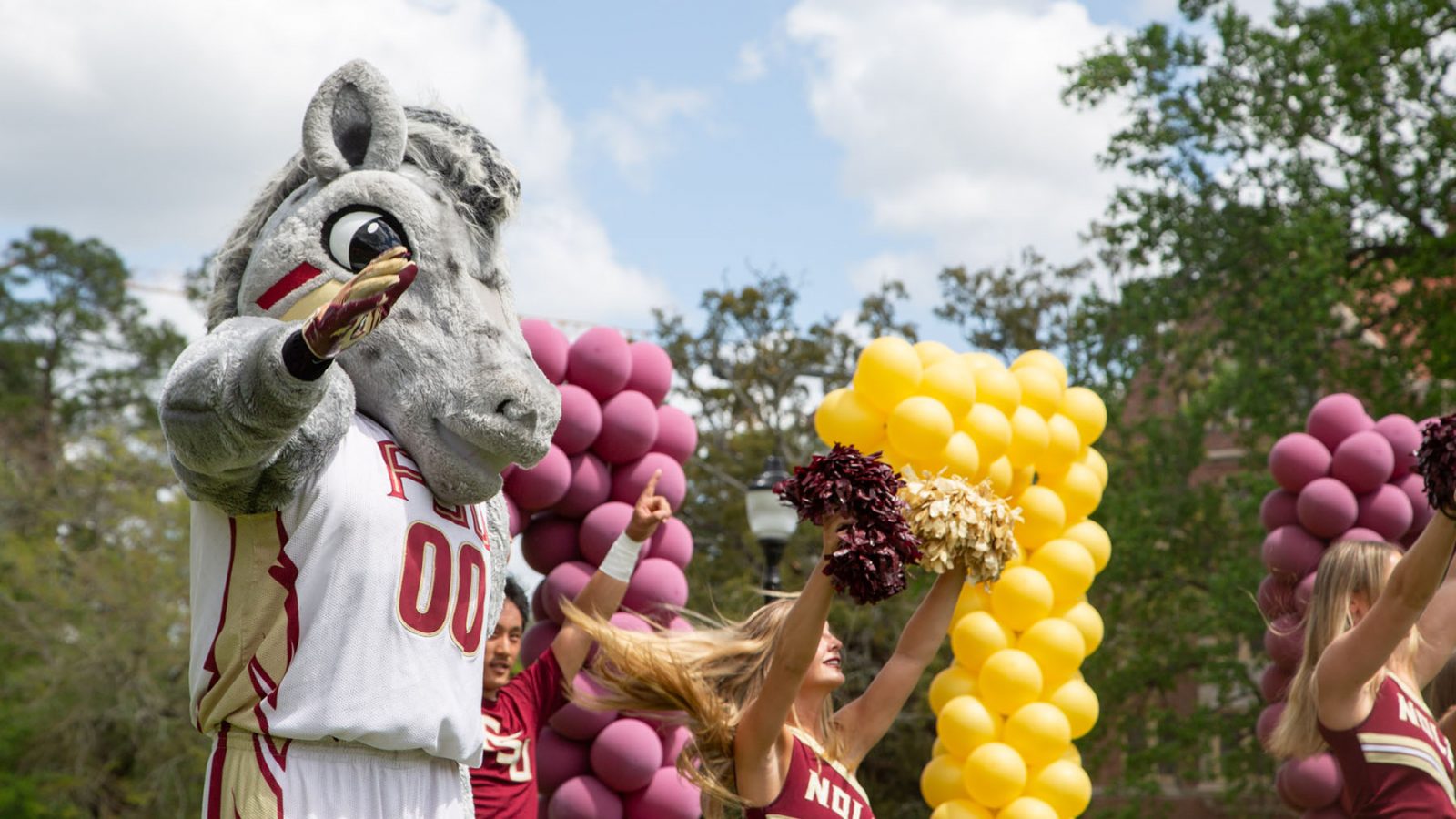 A mascot holds up his hand by cheerleaders and balloons, outside