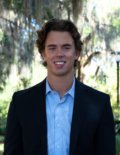 A professional headshot of a smiling young man with wavy brown hair, wearing a light blue button-down shirt and a black blazer, posing outdoors with sunlit Spanish moss in the background.