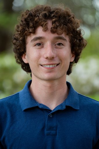 A young man with curly brown hair wearing a simple navy blue polo shirt.