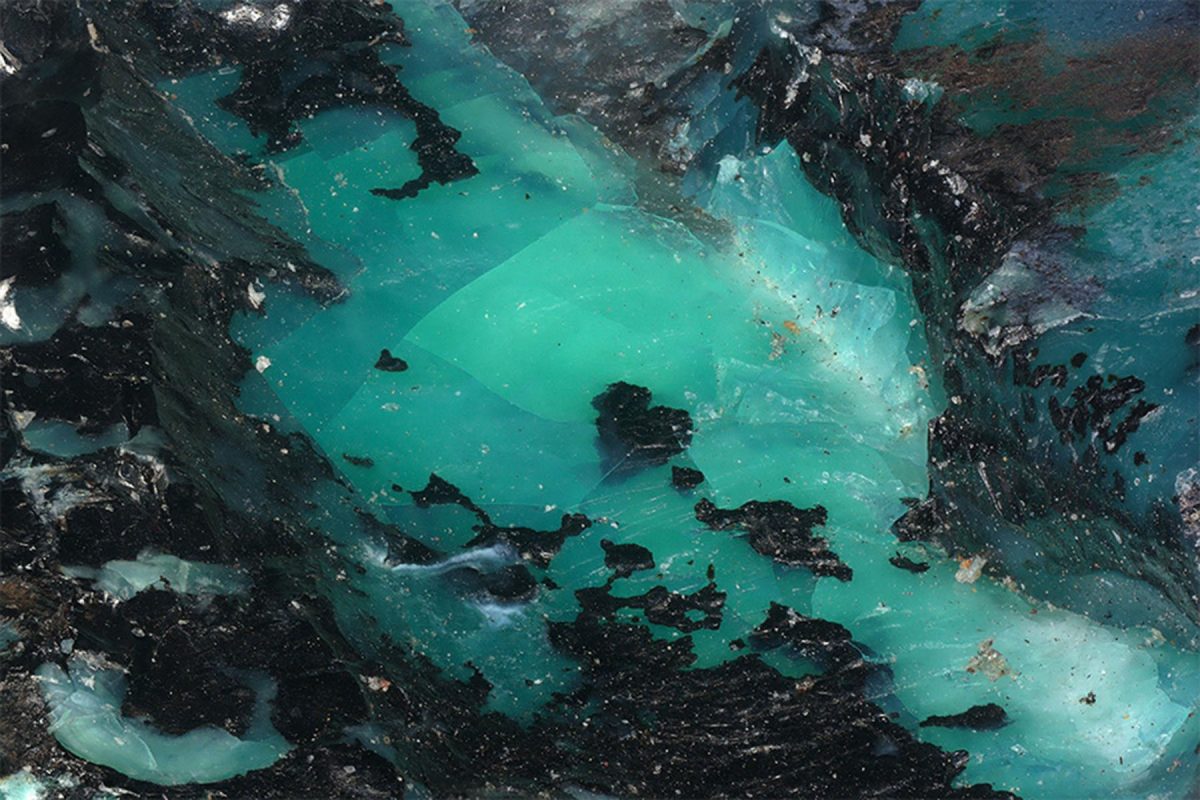 A vibrant, fractured core of translucent teal and seafoam green is encased in a jagged, dark obsidian-like border, creating a striking contrast of raw geological textures.