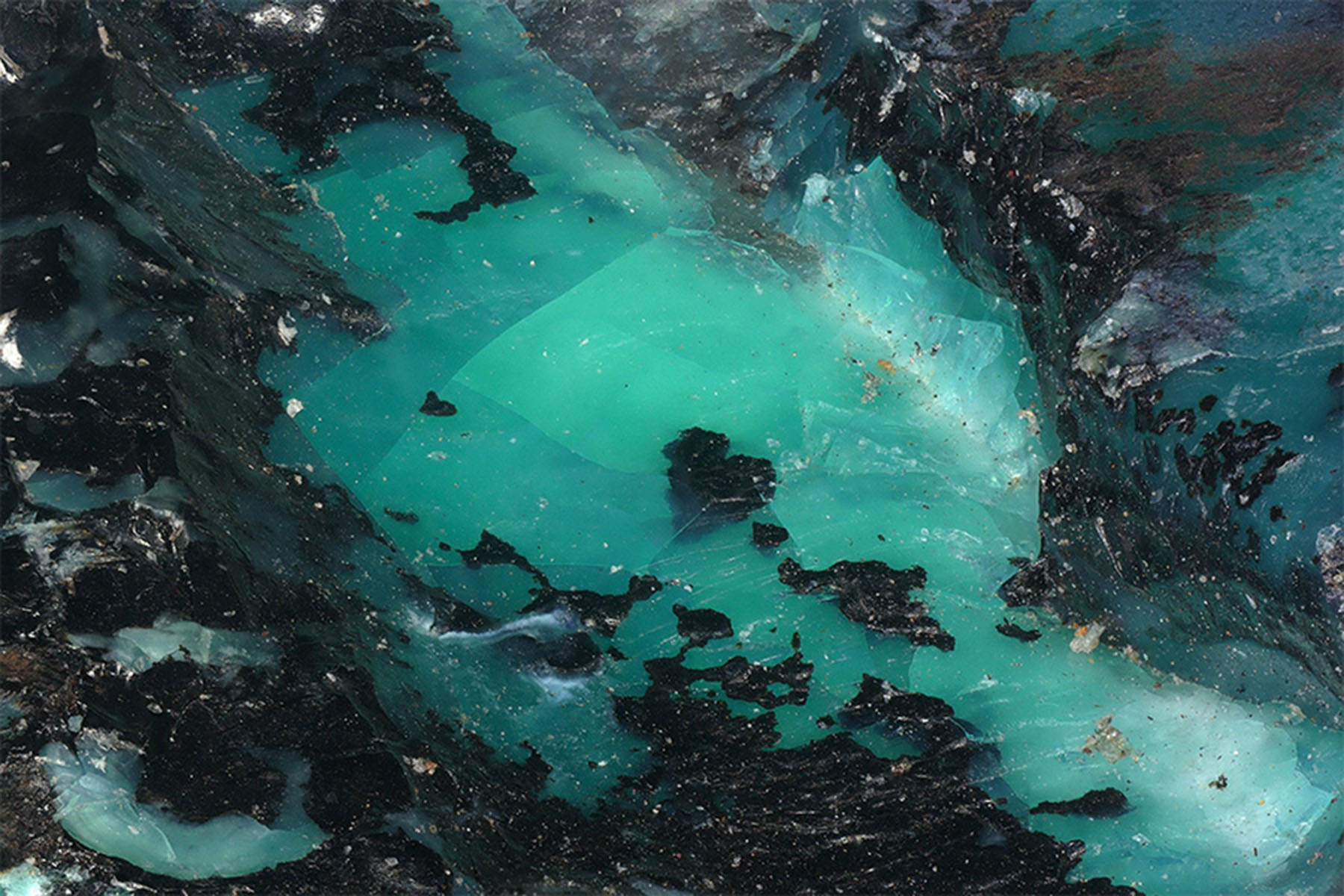 A vibrant, fractured core of translucent teal and seafoam green is encased in a jagged, dark obsidian-like border, creating a striking contrast of raw geological textures.